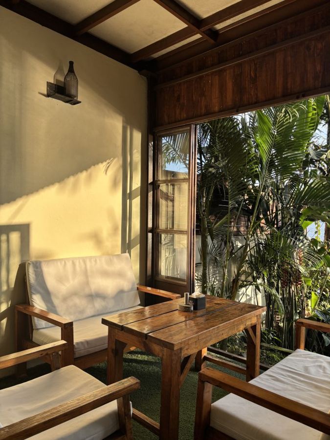 Varanda da cafeteria com mesa de madeira, poltronas claras e plantas ao fundo