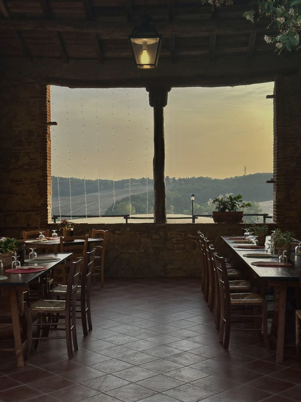 Salão do restaurante com mesas de madeira e janelas abertas para o pôr do sol